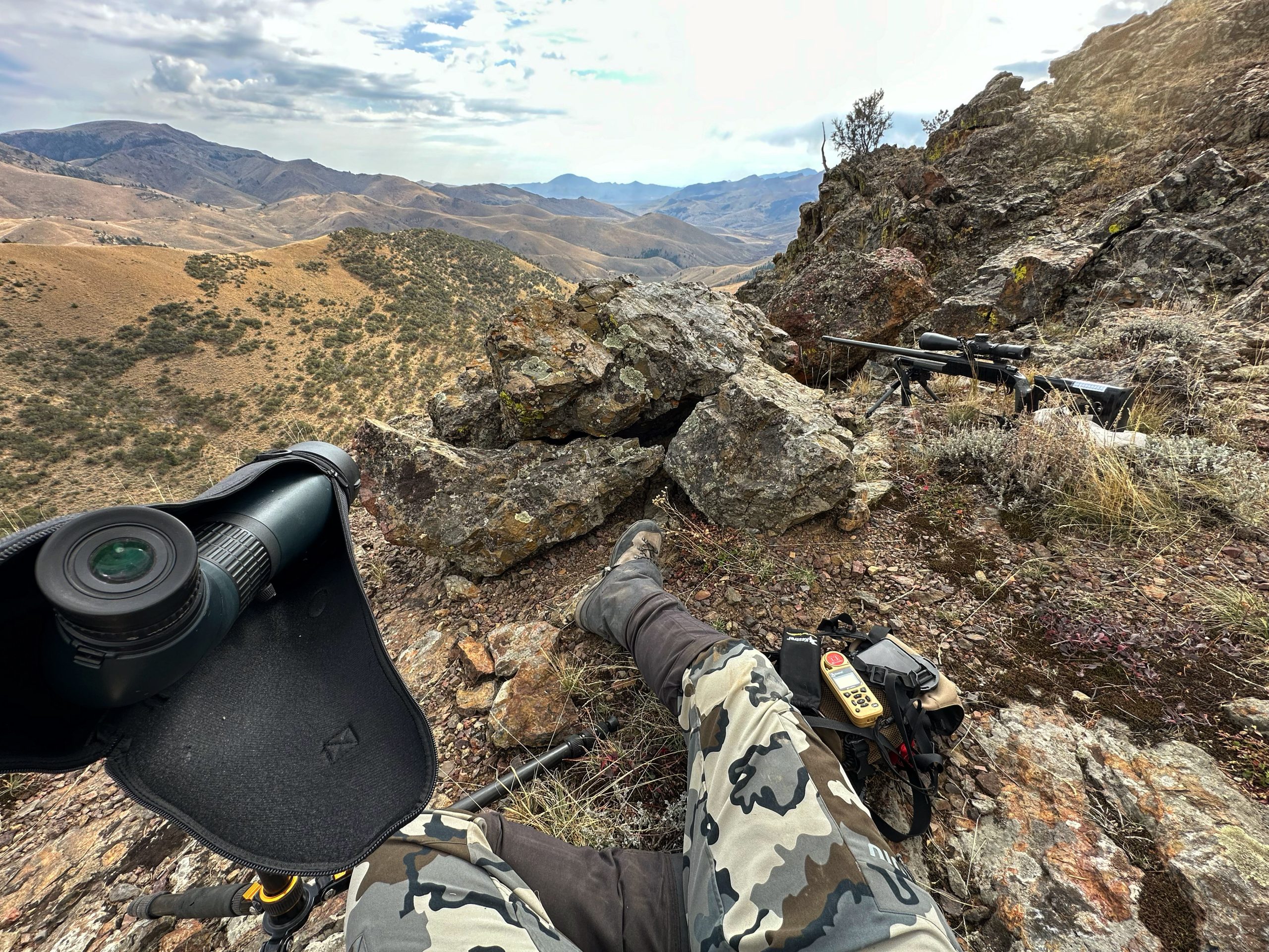 A hunter sits on top of a rocky outcropping, with his gun and spotting scope set up for a shot.