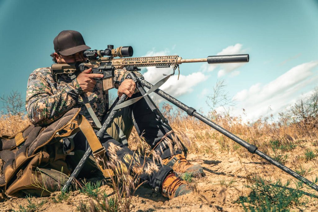 A shooter sitting in dry brush aiming through a rifle attached to a tripod