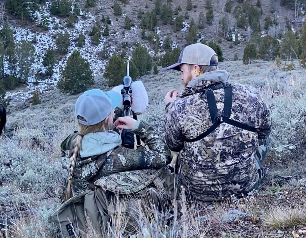 A hunter sets up for a shot at an animal while a second hunter walks her through what to do.