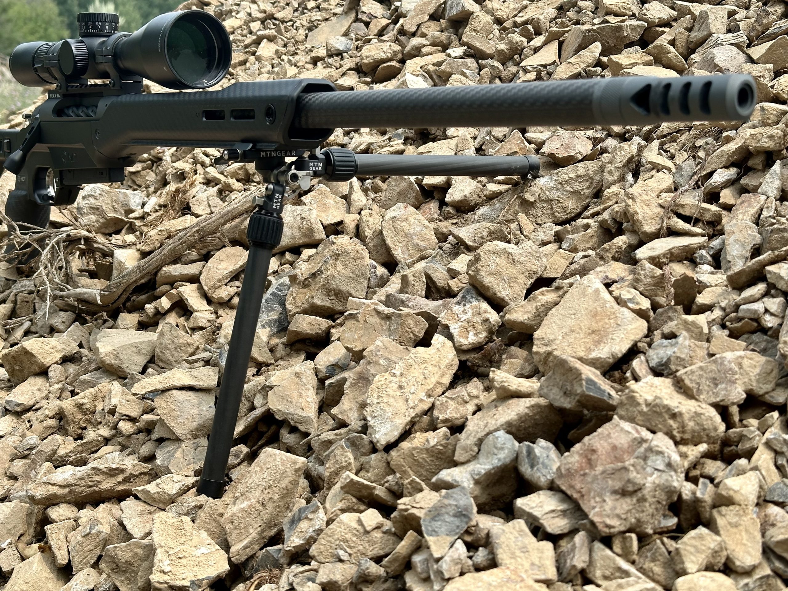 A black carbon fiber rifle is set up to shoot on a tan rock pile. 