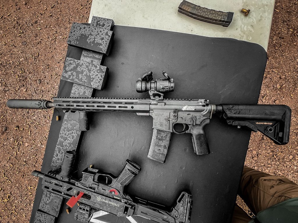 Among other rifles, there is a suppressed AR-style rifle laid on a table in the desert.