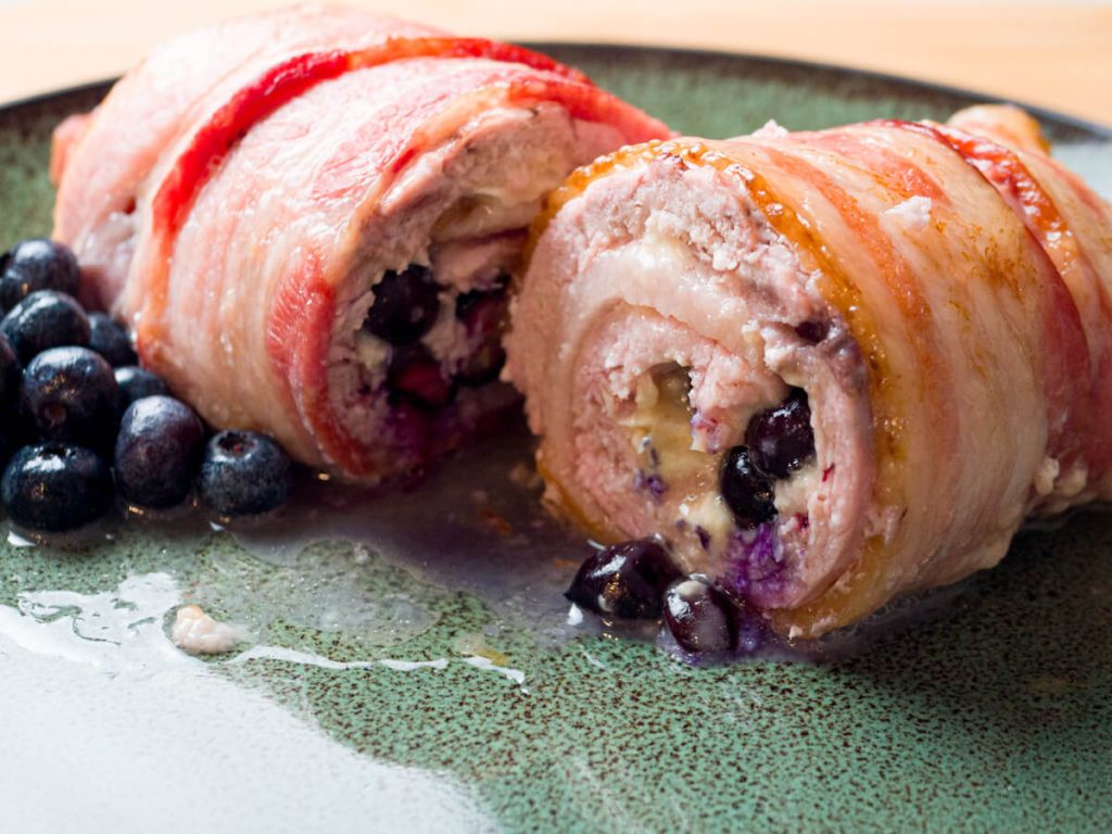 A roll of pheasant meat wrapped with bacon with blue berries and cheese inside is cut open revealing the berries and cheese. Blueberries garnish the plate.