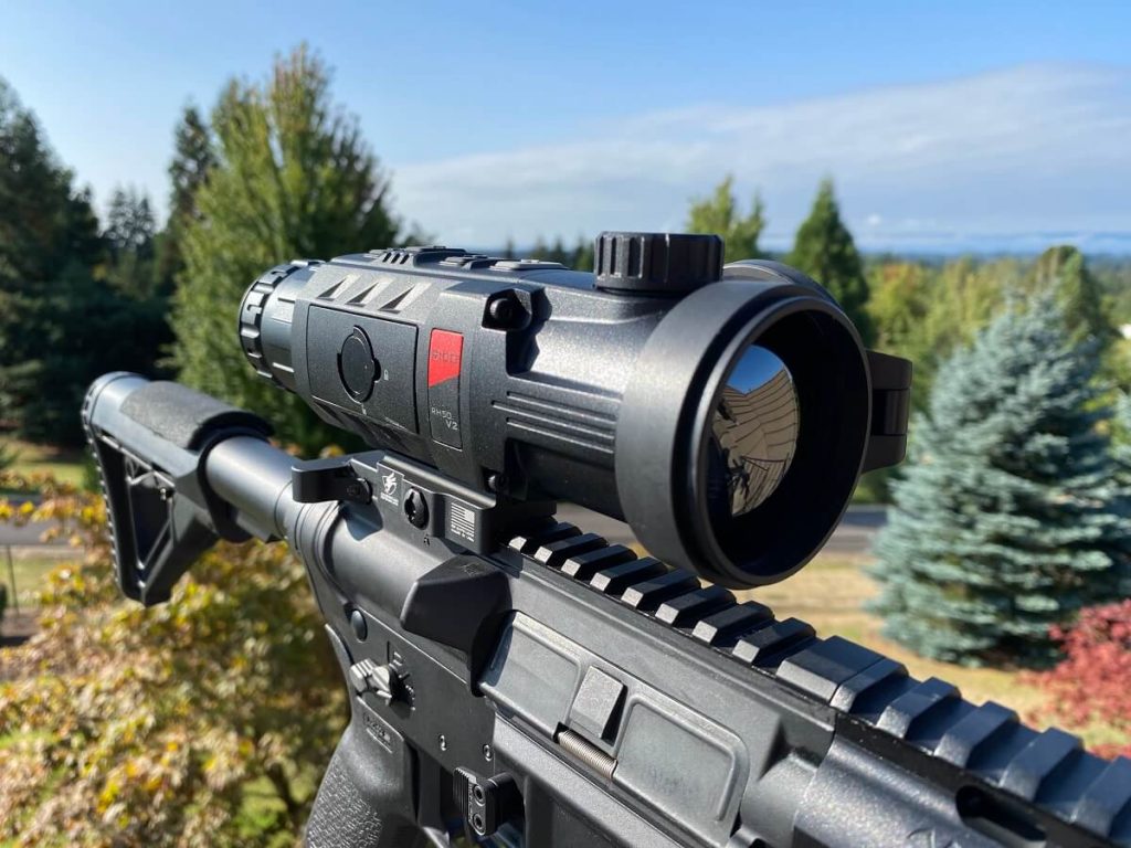 Riflescope mounted on AR-15 in front of backdrop of large evergreen trees and dry ground